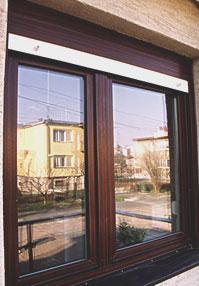 Brązowe okno PCV z roletą zewnętrzną i widokiem na budynek mieszkalny odbijający się w szybie.